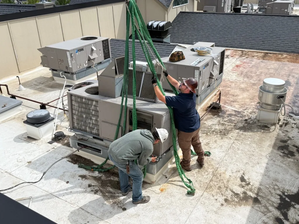 Two Unlimited techs in hard hats rigging a York package unit for removal on a flat commercial rooftop with green nylon slings and snow-capped mountains behind
