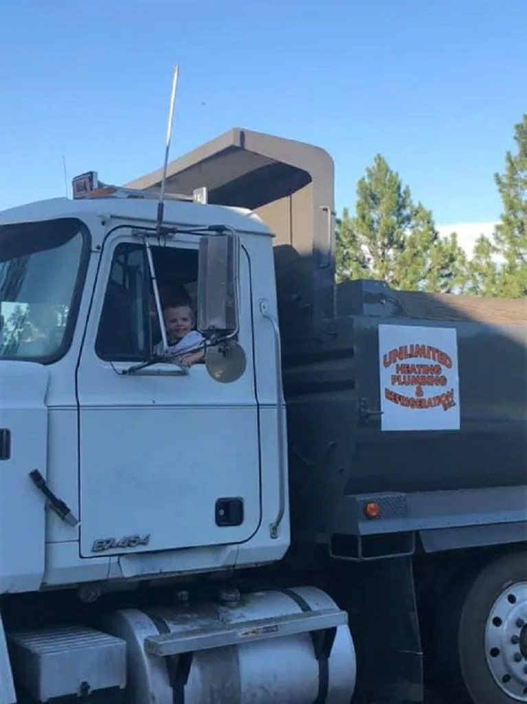 Young child in the cab of an Unlimited Heating, Plumbing & Refrigeration Mack tank truck under blue Palouse sky