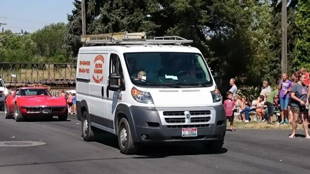 White Ram ProMaster Unlimited service van in a small-town Palouse parade, orange logo and phone numbers visible on the side