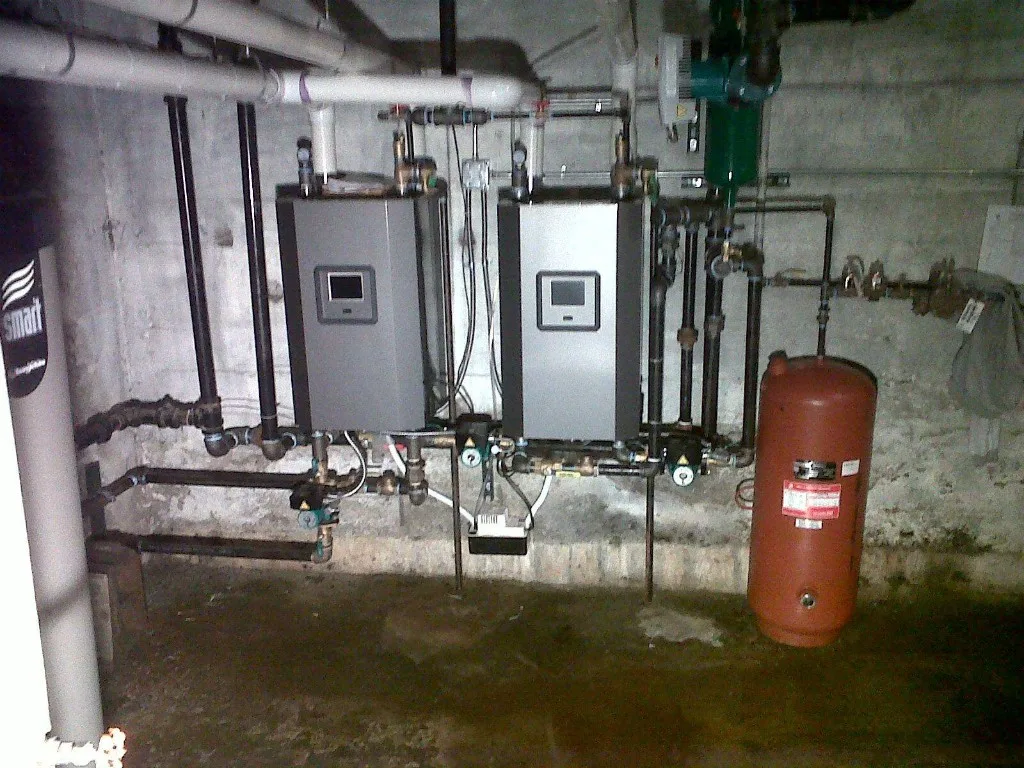 Pair of wall-hung hydronic boilers with a red expansion tank and smart controls in an unfinished basement mechanical room
