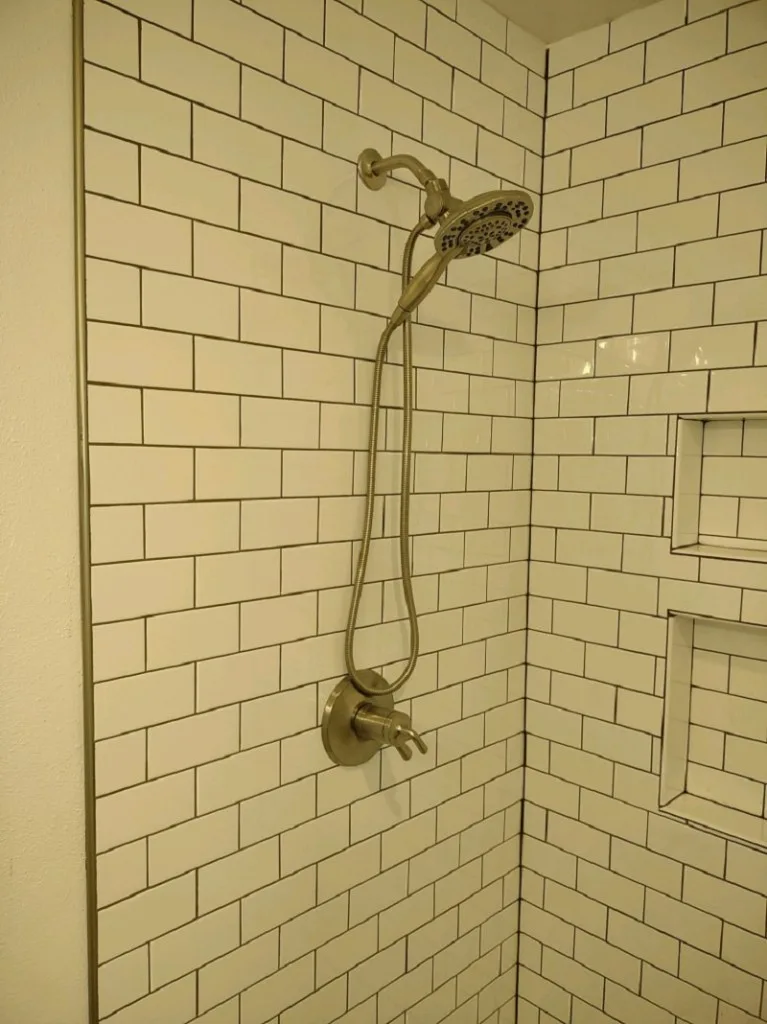 Brushed-nickel shower head and handheld wand on white subway tile with dark grout and a tile niche