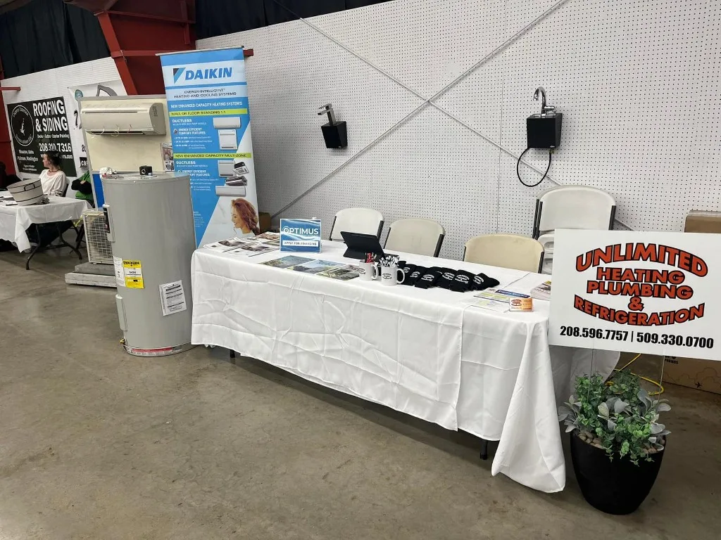 Unlimited trade-show booth with a Daikin banner backdrop, a ductless mini-split head on display, a Bradford White water heater, an Optimus 'Apply for Financing' placard, and a vertical sign reading 'Unlimited Heating Plumbing & Refrigeration'
